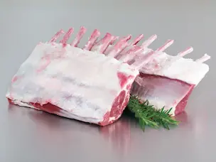 Raw rack of lamb with the fat cap removed, arranged on a clean surface with a sprig of herbs.
