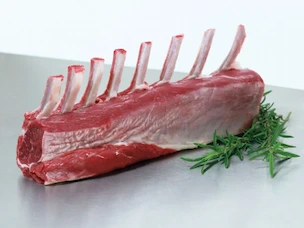 Raw frenched rack of lamb with exposed rib bones, placed on a metal surface with fresh herbs.